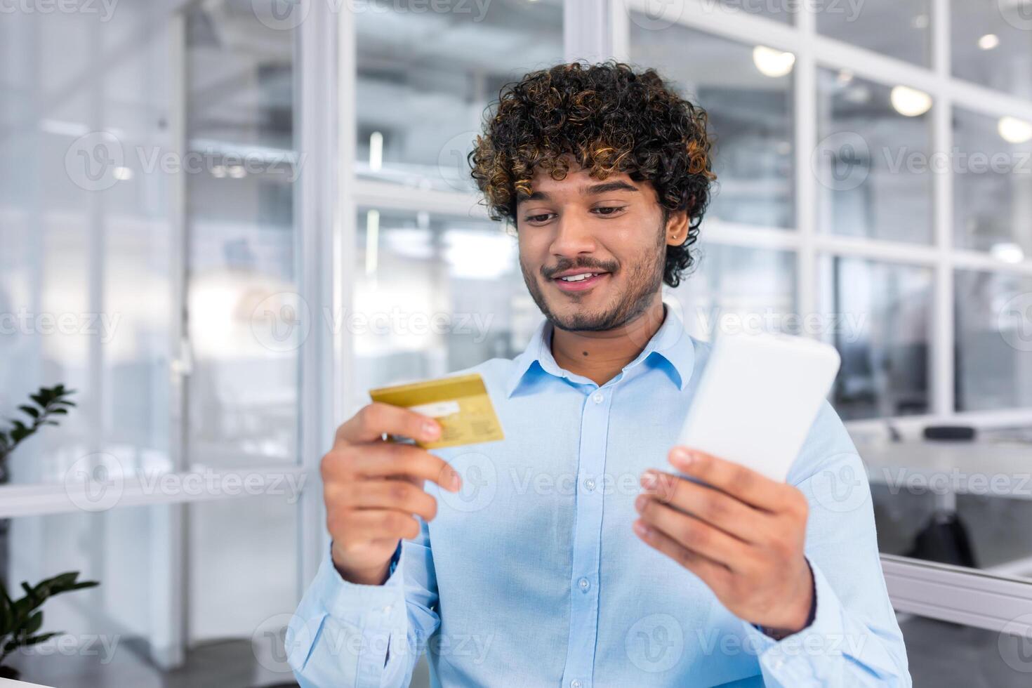Young successful indian man close up in modern light office interior holding bank credit card and phone, businessman smiling contentedly and making purchase in internet store online. photo