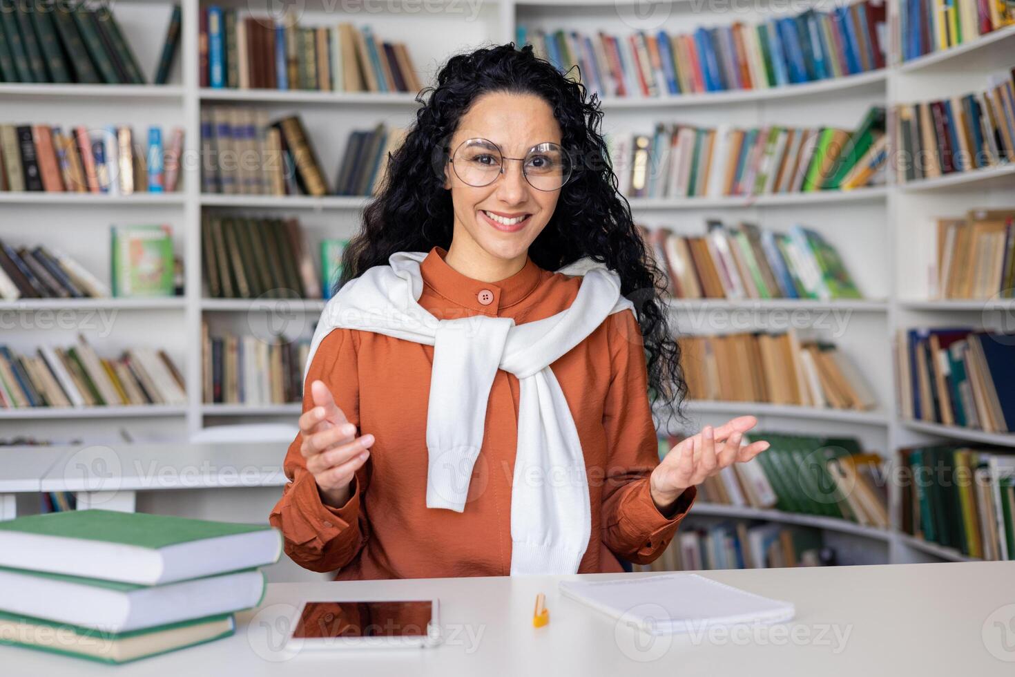 A young woman actively engaging with someone unseen, using expressive hand gestures, sitting in front of a bookshelf with books. photo