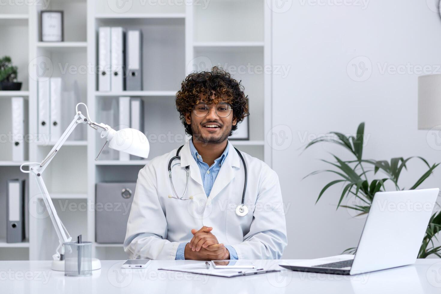 Portrait of young Indian doctor, man in white medical coat smiling and looking at camera, doctor sitting at table inside medical office of clinic, working with laptop. photo