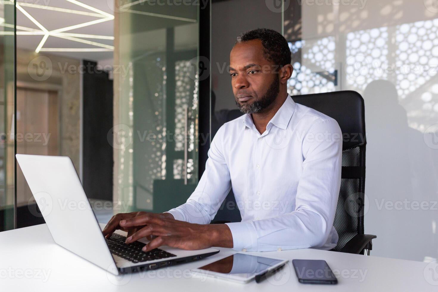 Serious focused african american programmer working inside office, man coding new software on laptop, mature experienced team leader developing technical project. photo