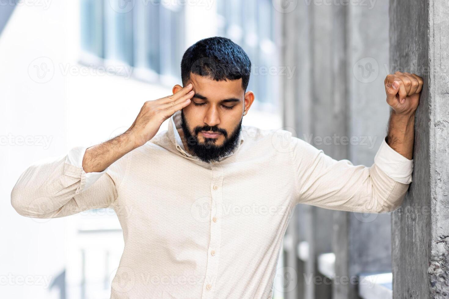 Close-up photo of a young Arab man in a shirt and jeans standing outside with his eyes closed, leaning against the wall of a building and holding his head. Feels strong pressure and pain