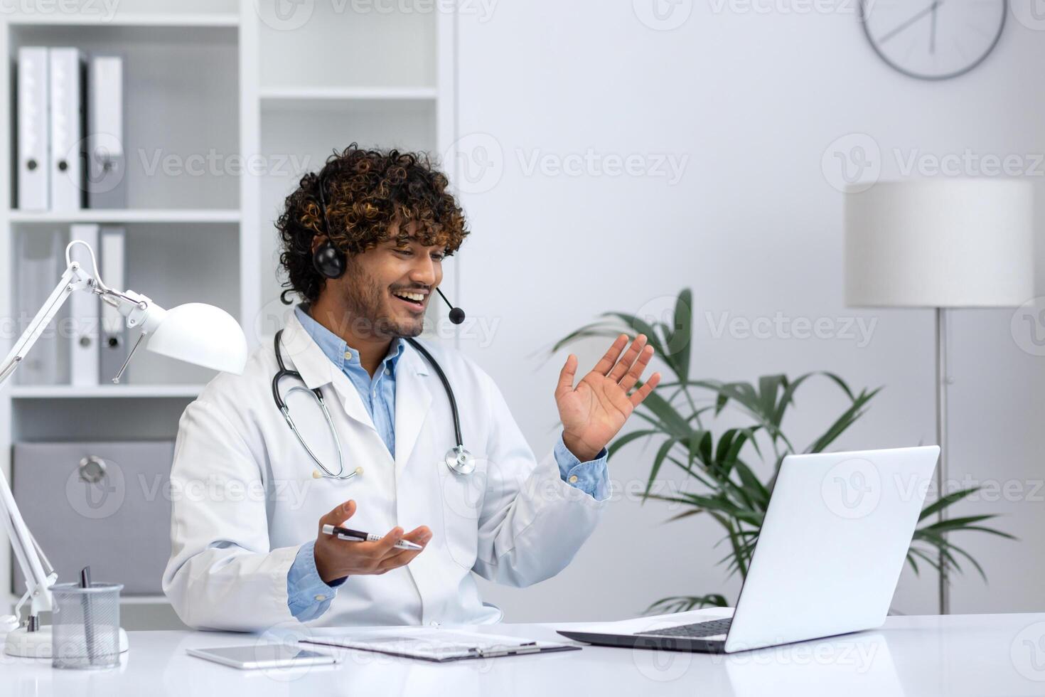 Young Indian doctor consulting patients remotely, man in white medical coat with headset using laptop for call, smiling contentedly inside clinic office. photo