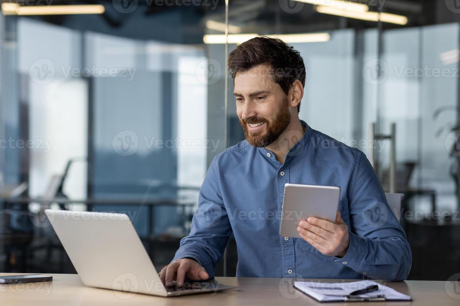 Smiling young businessman working on a laptop in the office, sitting at a desk, typing on a keyboard, using a tablet. photo