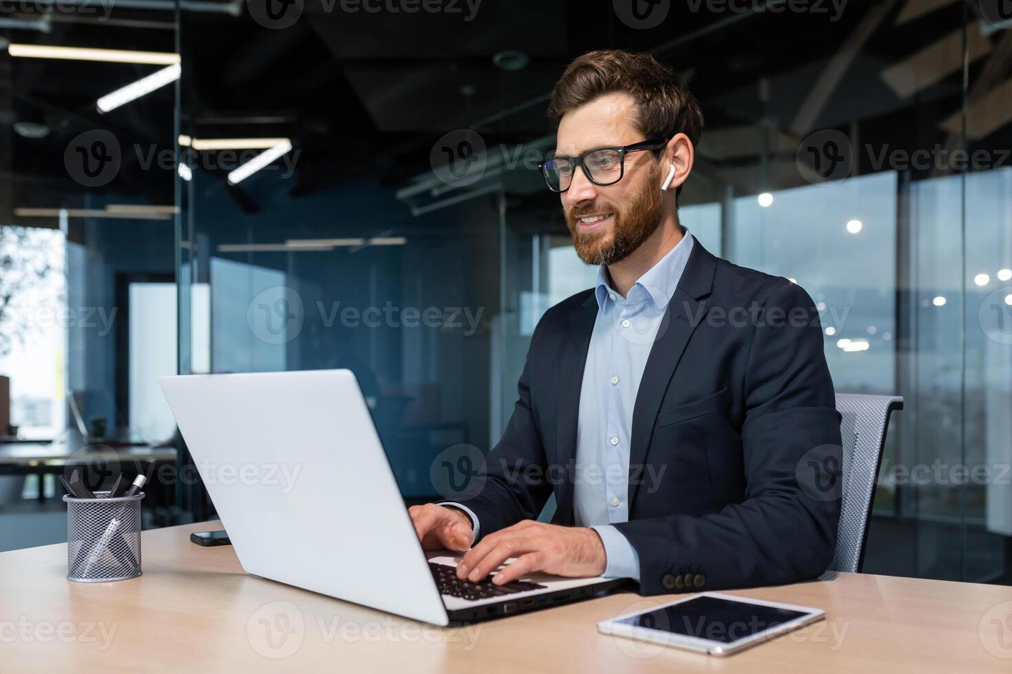 Mature businessman in headphones small earbuds talking on a call using a laptop, boss at work at the desk in a business suit in the middle of the office smiling friendly. photo