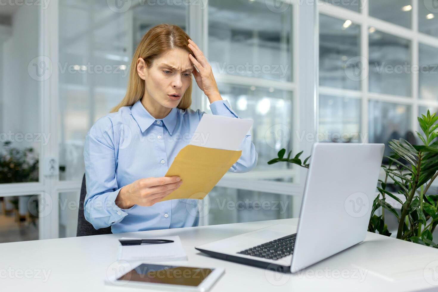A businesswoman in a blue shirt appears perplexed analyzing documents beside her laptop in a bright office setting. photo