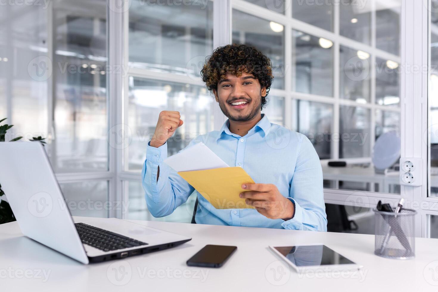 A young man, an Indian student, received good news by letter, he entered the university, passed the exam, and got a job. Sitting in the office, happy, smiling, looking at the camera. photo