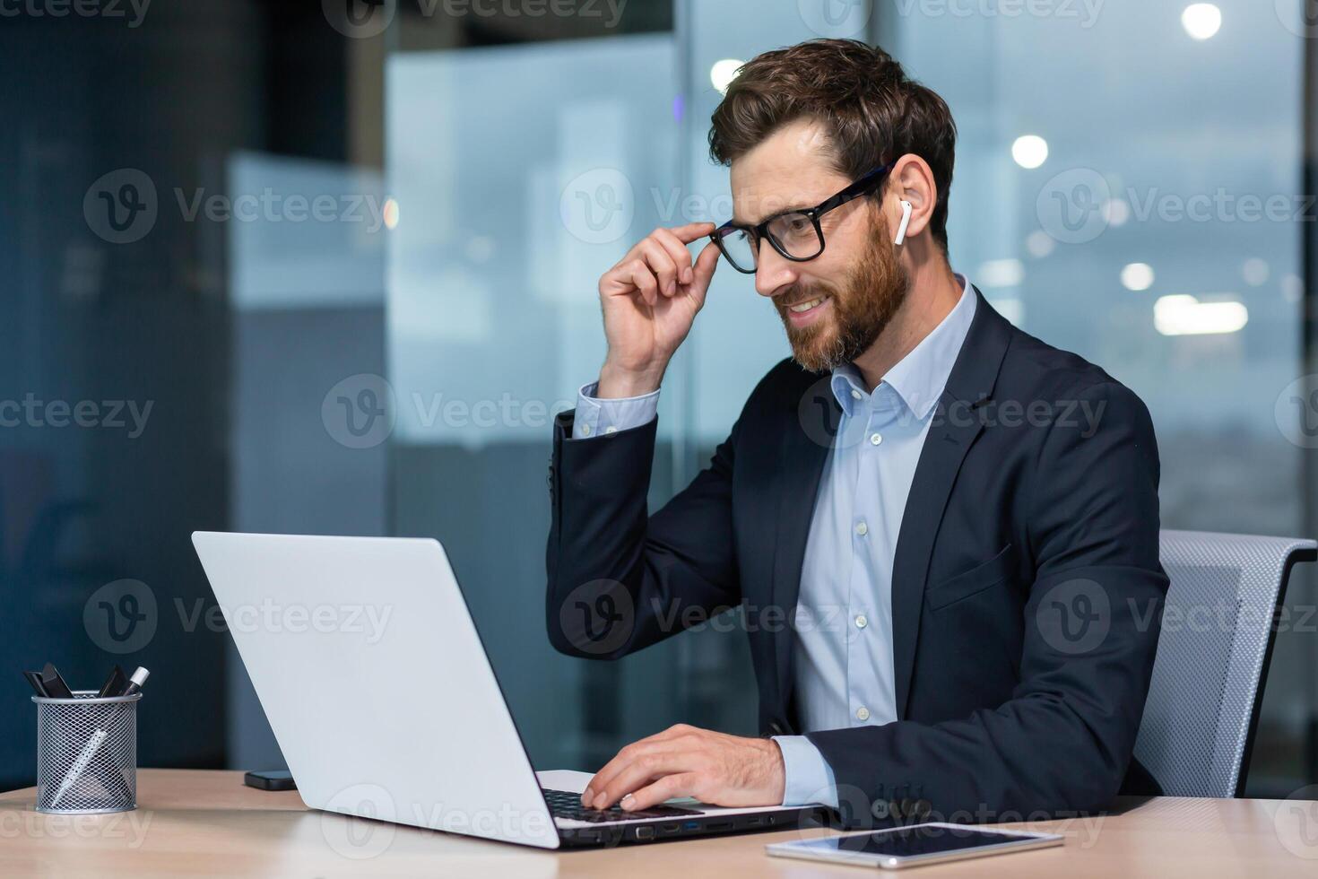 maduro empresario en auriculares pequeño auriculares hablando en un llamada utilizando un computadora portátil, jefe a trabajo a el escritorio en un negocio traje en el medio de el oficina sonriente amigable. foto