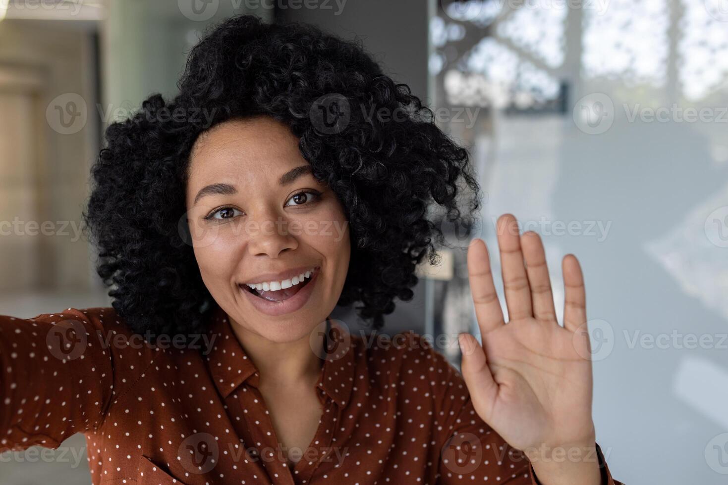 Beautiful business woman with curly hair wearing looking at smartphone camera, waving her hand in greeting gesture, talking with friends and colleagues, using call app photo