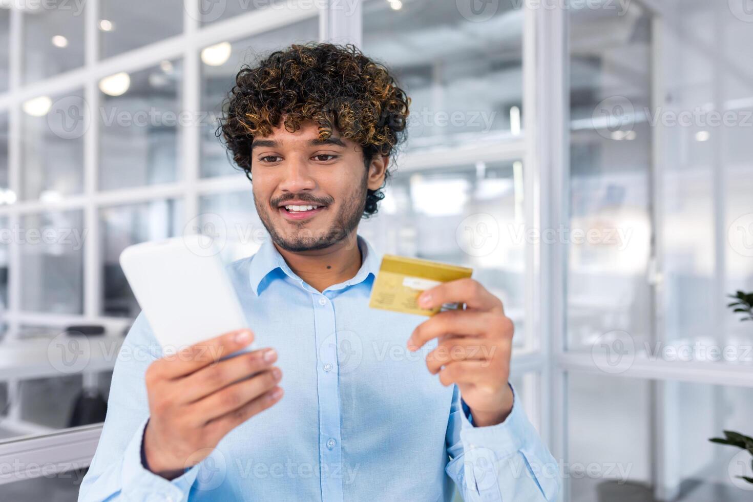 Young successful indian man close up in modern light office interior holding bank credit card and phone, businessman smiling contentedly and making purchase in internet store online. photo