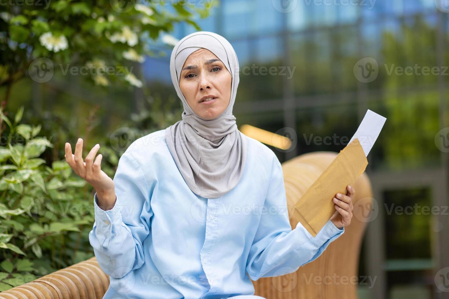 A woman in a hijab appears puzzled with an envelope in her hand, standing outside a modern building. photo