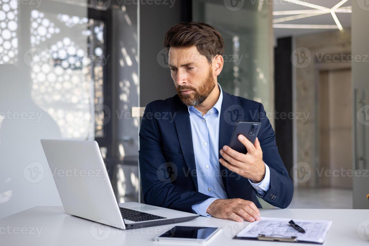 A serious young man businessman sits in the office at the table in a suit, works on a laptop and holds a mobile phone in his hands. photo