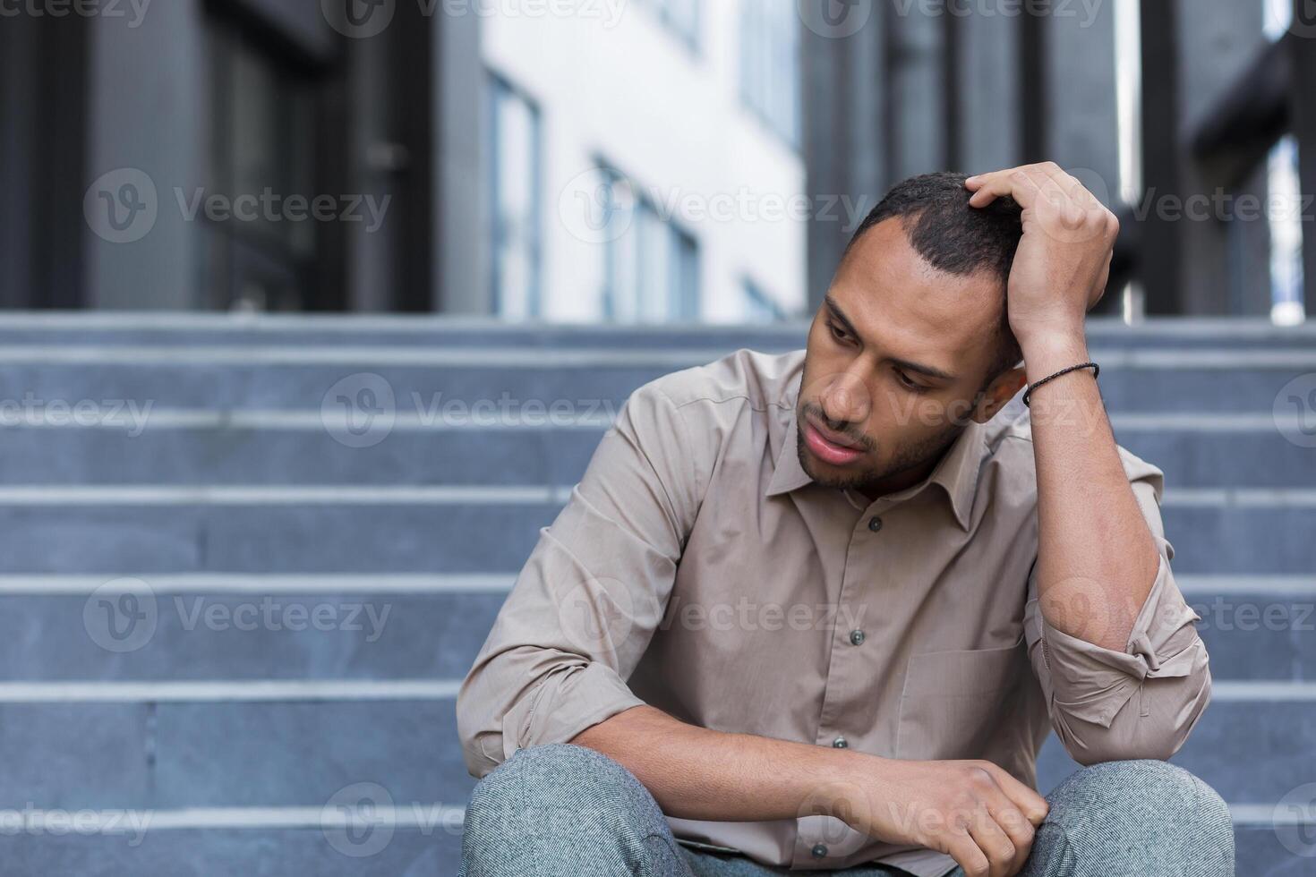Disappointed and sad man sitting on stairs outside office building, employee fired from work, african american man in shirt upset and depressed photo