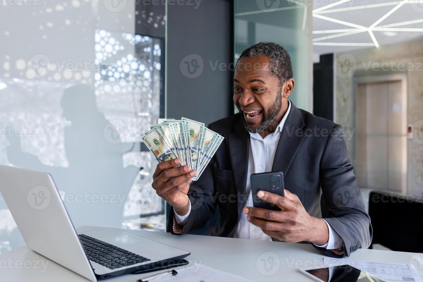 Happy African-American man sitting at the desk in the office, holding a mobile phone in his hand and looking enthusiastically at a fan of cash money. photo