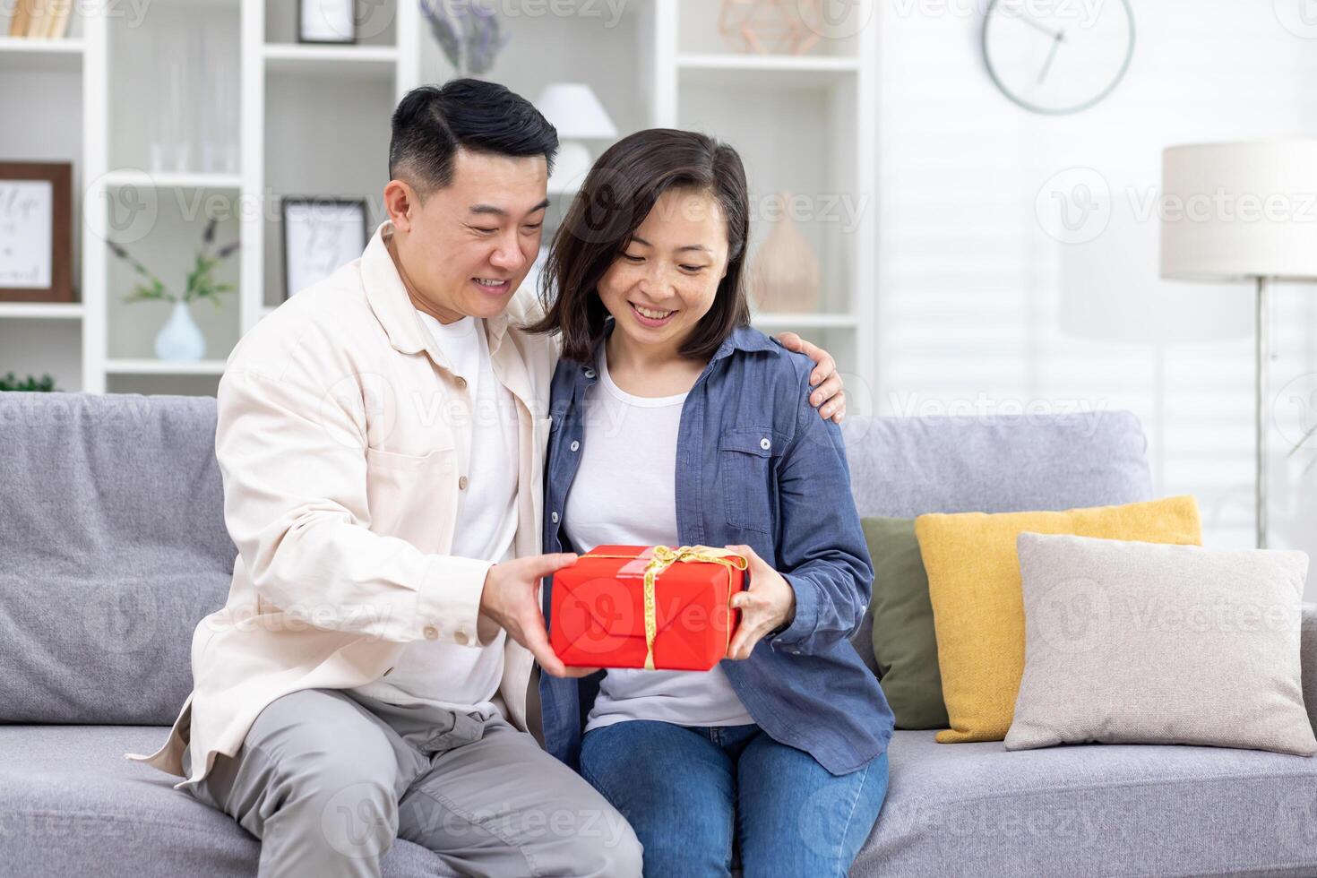 Happy young Asian couple, man and woman sitting on sofa at home. A man gives a woman a red box as a surprise gift. They smile at each other. photo