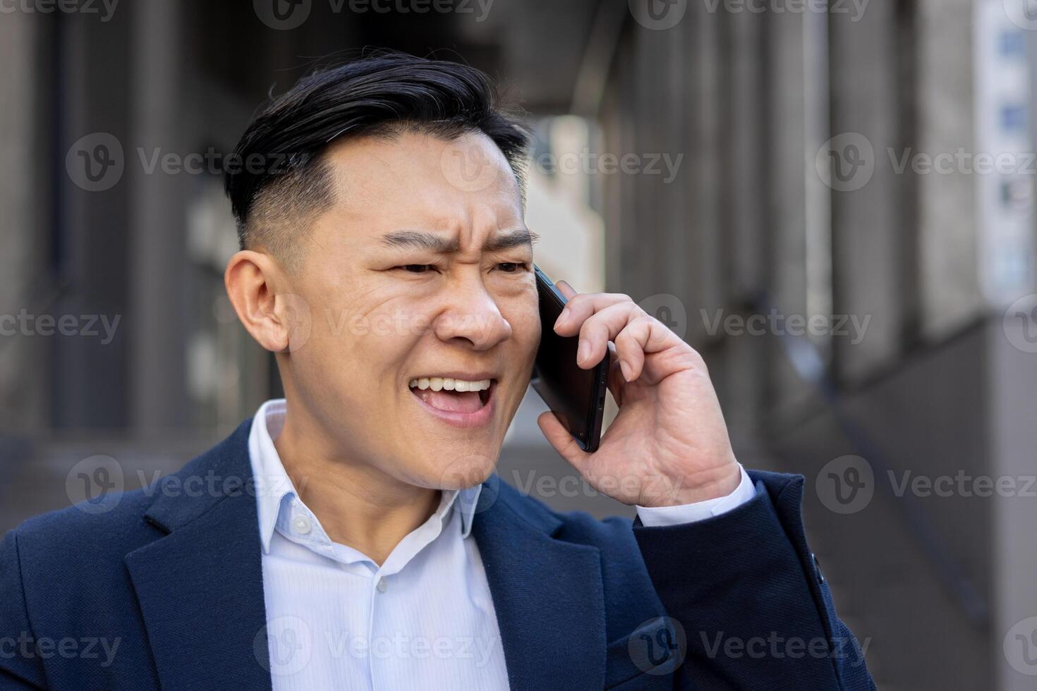 Annoyed Asian adult male in a business suit on a call outdoors, showing signs of frustration and urgency. photo