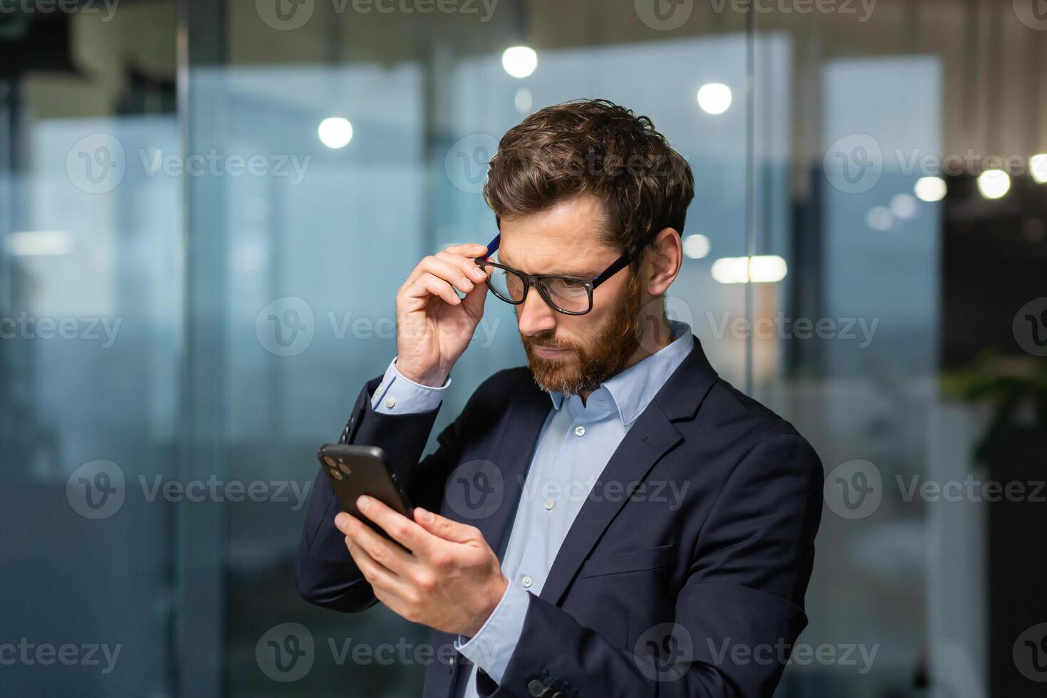 maduro empresario en oficina cerca ventana en noche trastornado leyendo mensaje en línea notificación desde teléfono, masculino jefe trabajando dentro oficina en negocio traje y lentes con barba, trastornado inversor. foto