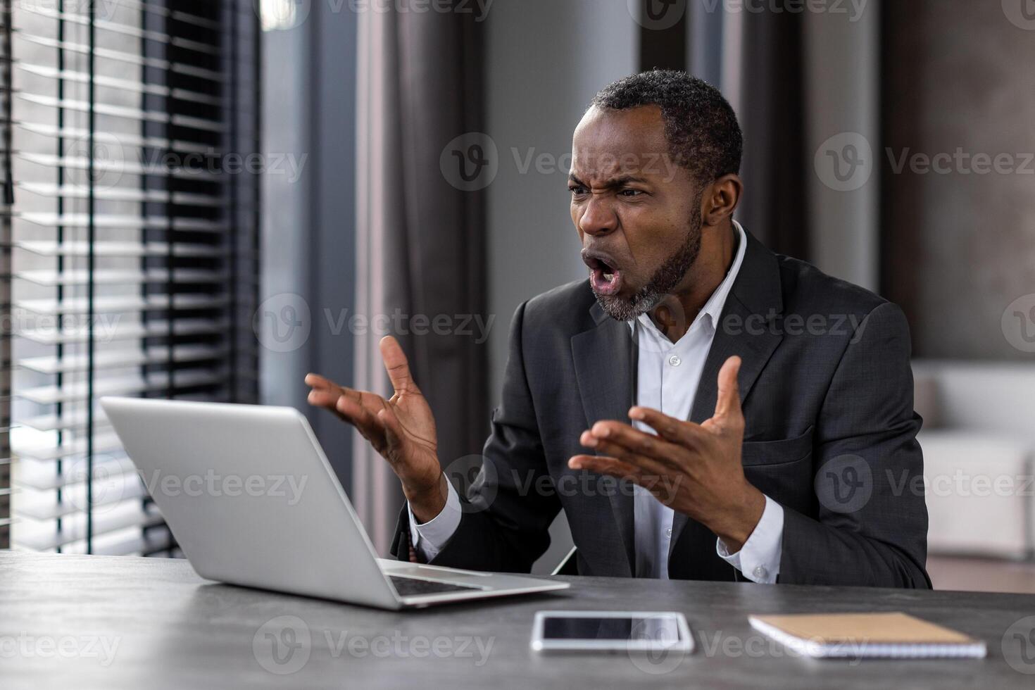Aggressive black man screaming in despair while working by laptop in stylish apartment interior. African american male worker shocked by computer operational breakdown and system error on device. photo