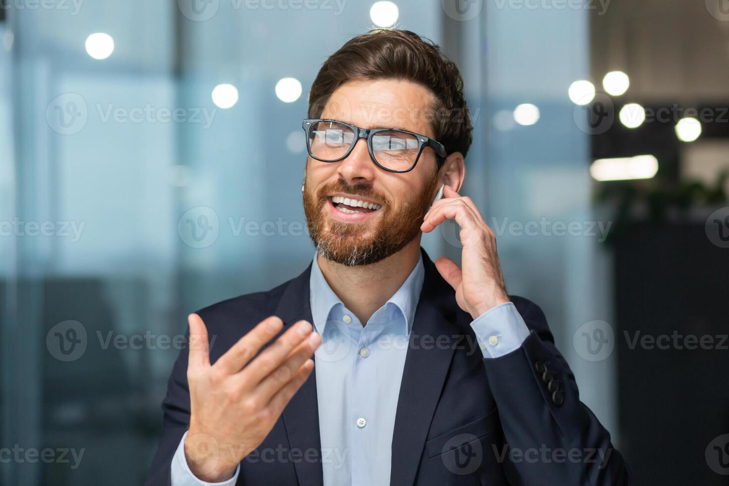 Close-up photo. A young man in the office and talking through a headset through his hands free. He is holding an earphone with his hand, gesturing with his hand, smiling. photo