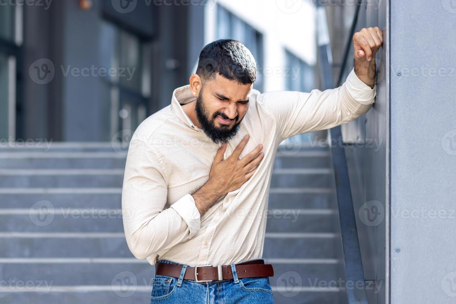 Panic and heart attack on the street. A young Indian man is standing near a building, leaning against a wall and holding his chest with his hand, feeling severe pain and needing medical help photo