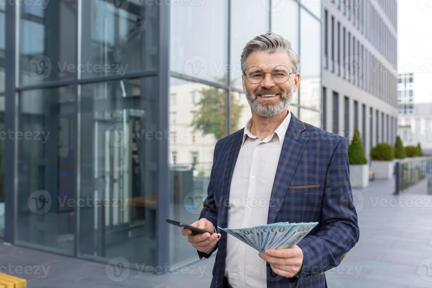 Happy boss standing on the street with a stack of dollars in his hands and a phone, winning the lottery, success at work, smiling and looking at the camera. photo