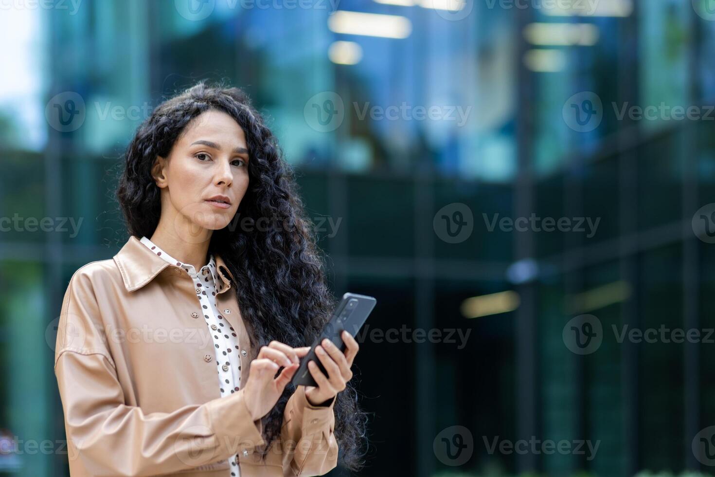 Serious mature woman boss outside office building looking thoughtfully at camera business woman holding phone in hands using app on smartphone, Hispanic woman with curly hair browsing internet pages photo