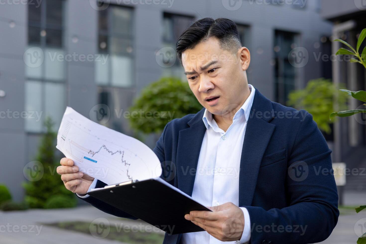 Worried and serious young Asian businessman standing outside office center in suit, holding documents and looking at graphs in frustration. photo