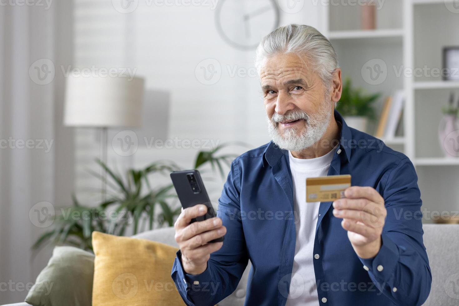 Smiling elderly man holding a credit card and using a mobile phone in a cozy living room setting, exemplifying online shopping or banking. photo