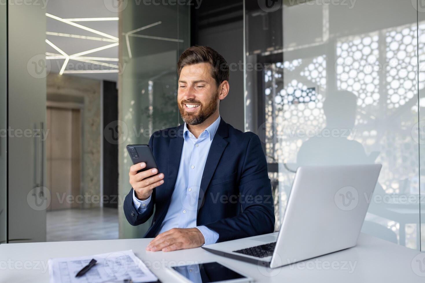 A relaxed and smiling young man in a business suit sits in a chair at a desk in the office and looks at the screen of the phone, he is holding in his hands. photo