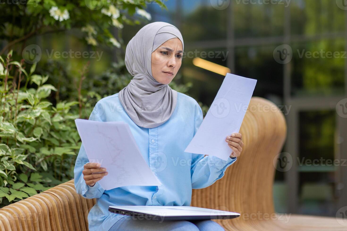 A focused professional woman in a hijab examines papers, sitting on an outdoor bench in a tranquil setting. photo