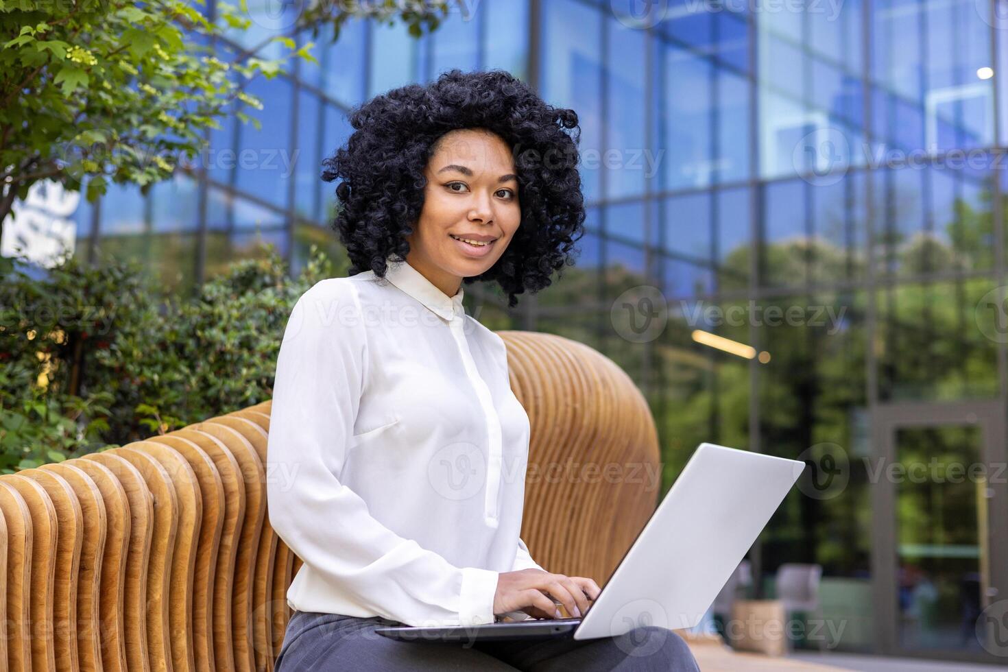 retrato de un joven africano americano mujer de negocios sentado fuera de un oficina centrar en un banco y utilizando un ordenador portátil. él sonrisas y mira con aire de suficiencia dentro el cámara. foto