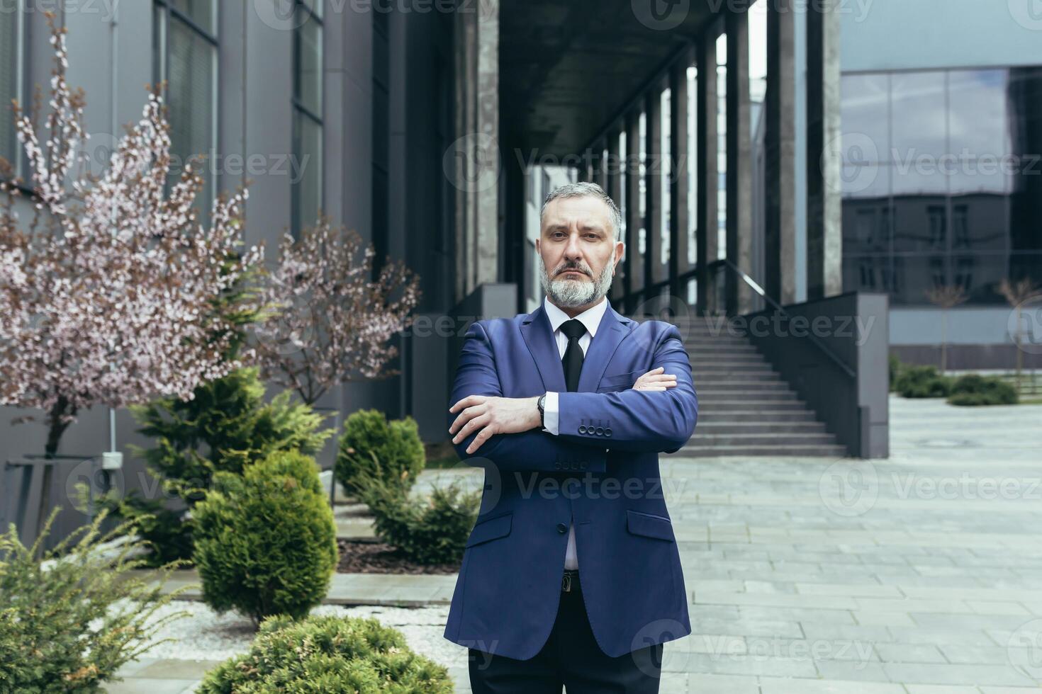 Portrait of senior handsome man, a businessman, a judge, a lawyer. Standing in a suit near the courthouse, office center. Crossed his arms in front, looks at the camera, serious. photo