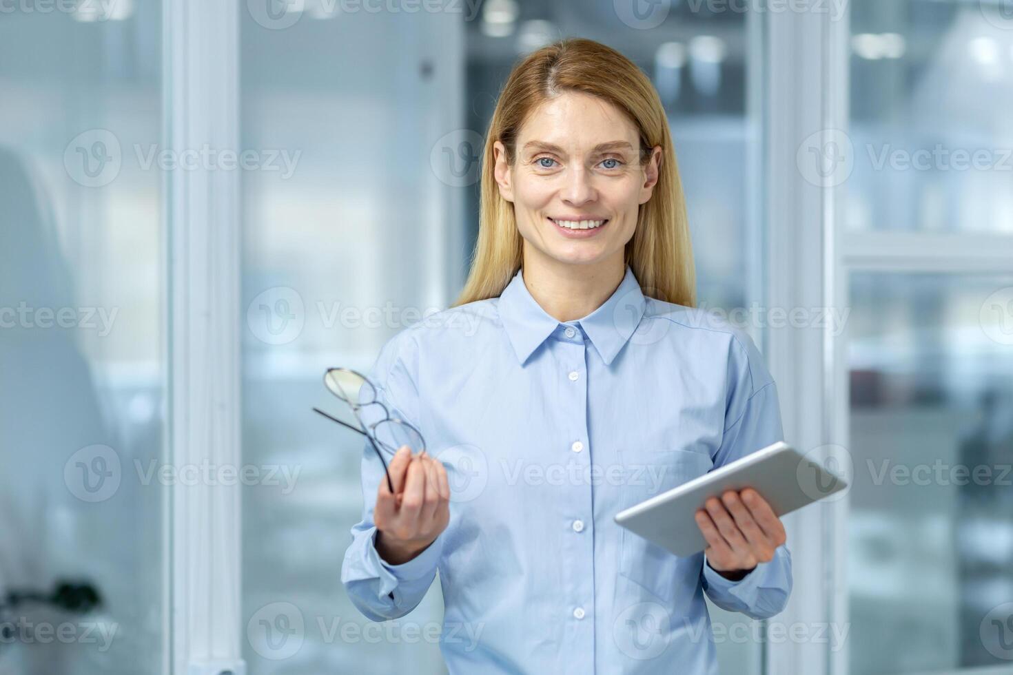 hermosa mujer de negocios sonriente a cámara con elegante los anteojos y electrónico tableta en manos. medio años confidente empresario consiguiendo Listo para nuevo trabajando día en elegante corporativo edificio. foto