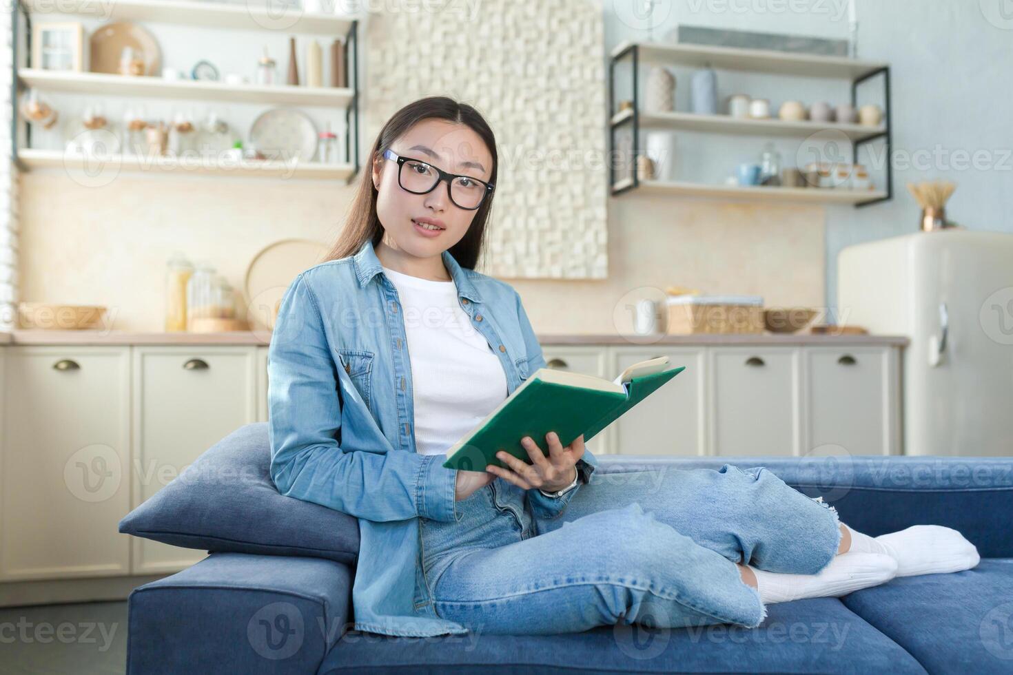 Portrait of a young beautiful Asian woman in denim clothes and glasses sitting at home on the sofa on the background of the kitchen. He holds a green book in his hands, reads, looks at the camera. photo