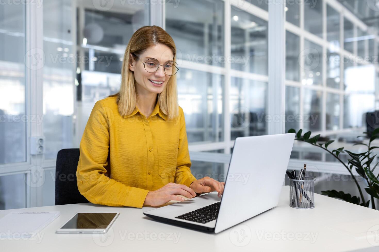 Happy and successful business woman working inside office with laptop, female worker in yellow shirt and glasses typing on keyboard, woman satisfied with work and happy at workplace sitting at table. photo