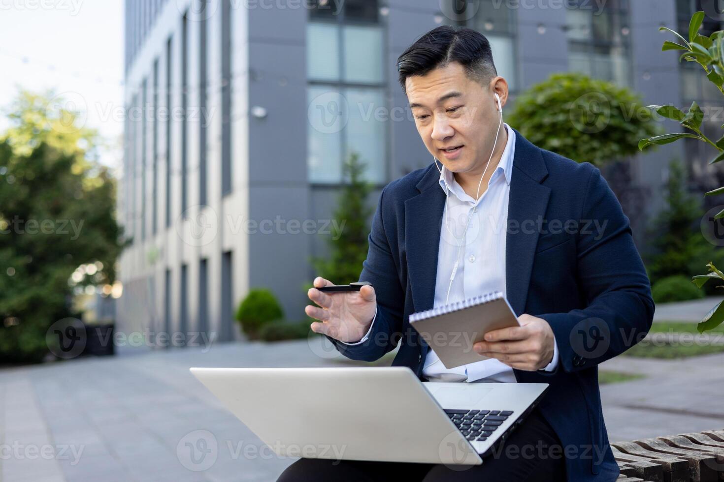 asiático joven empresario sentado cerca oficina edificio en un banco vistiendo auriculares, hablando en un llamada mediante un ordenador portátil y haciendo notas en un cuaderno. foto