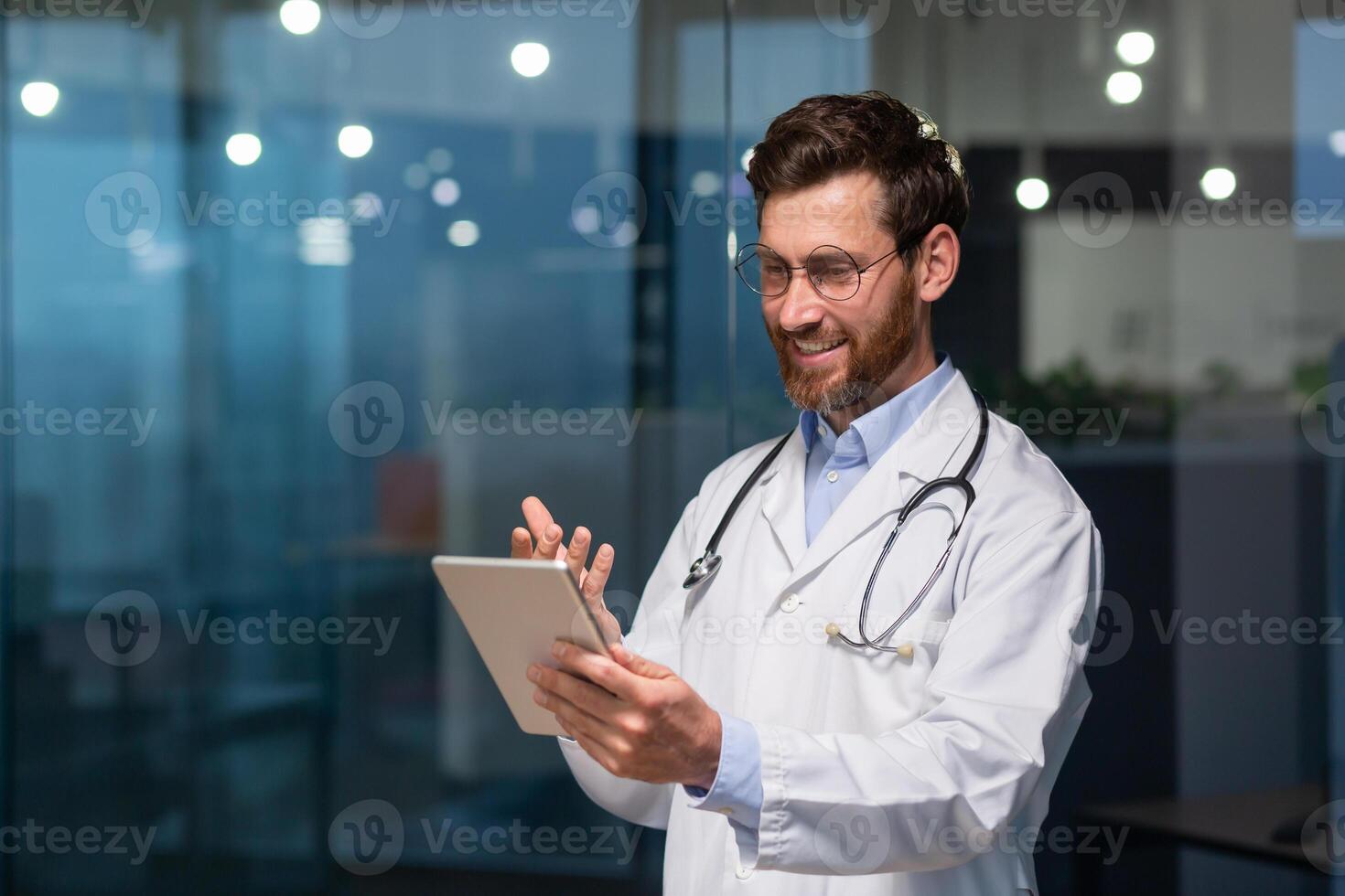 A young male doctor is talking to a patient through a call from a tablet. Standing in the office in the clinic in a white coat and with a stethoscope photo