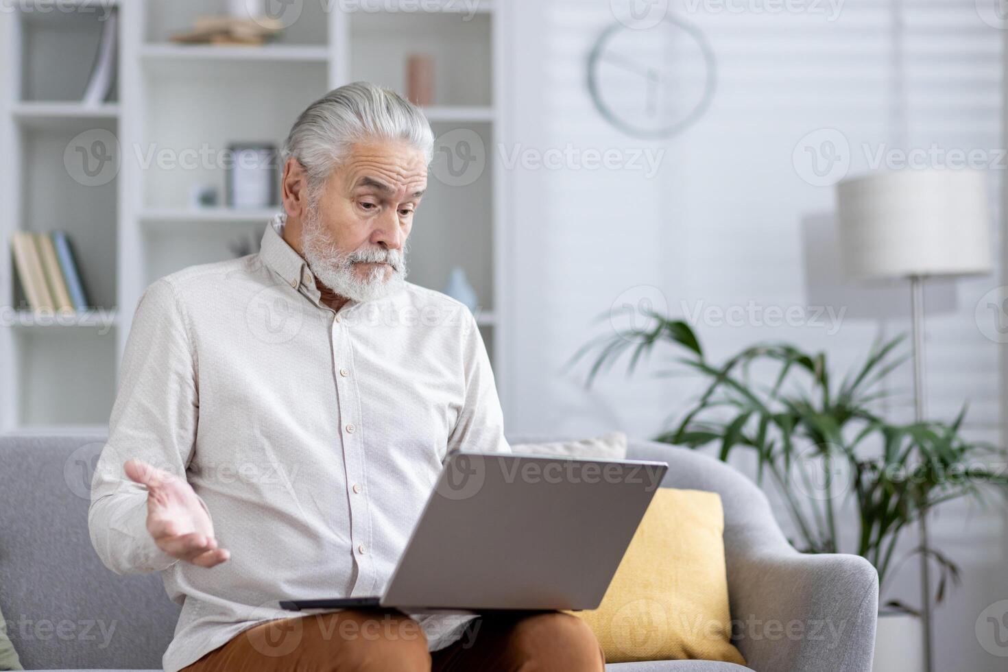 Focused senior male with a beard in a bright room, looking puzzled at a laptop while sitting on a couch. photo