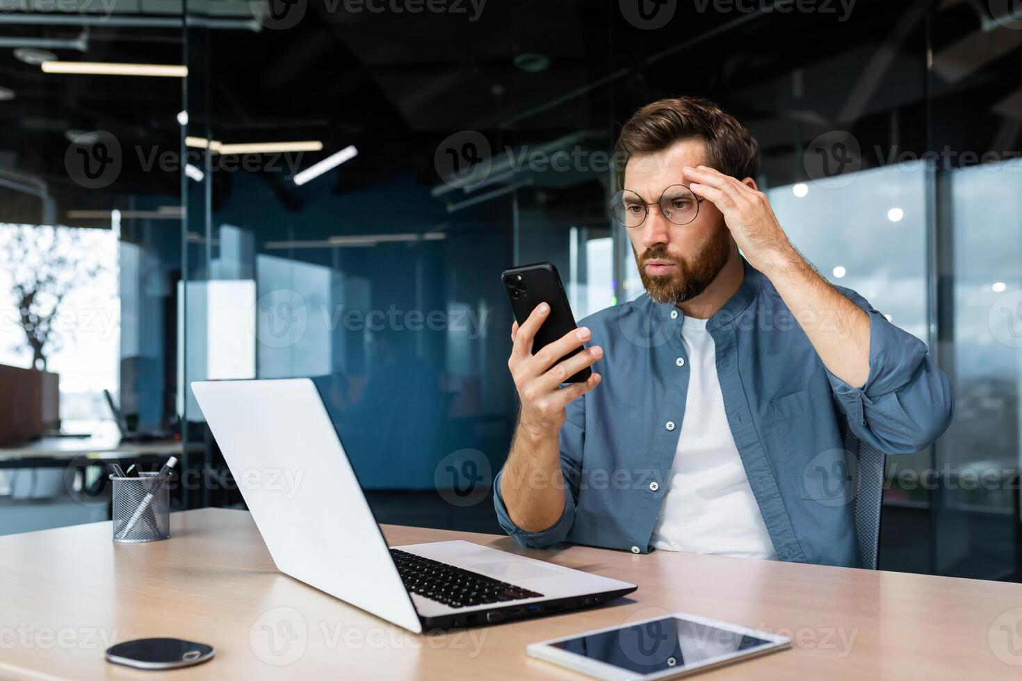 Serious mature boss working inside modern office building with laptop, businessman received bad news online, reading on smartphone confused angry and depressed man at work. photo