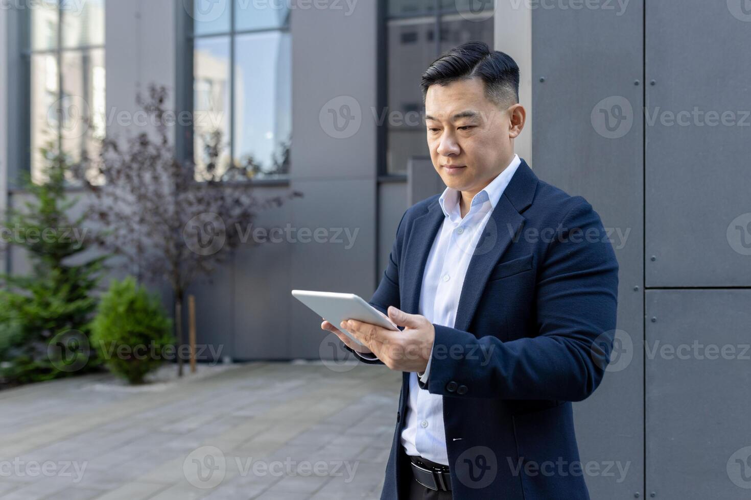 An Asian businessman in a suit stands outside an office, focused intently on his digital tablet. photo