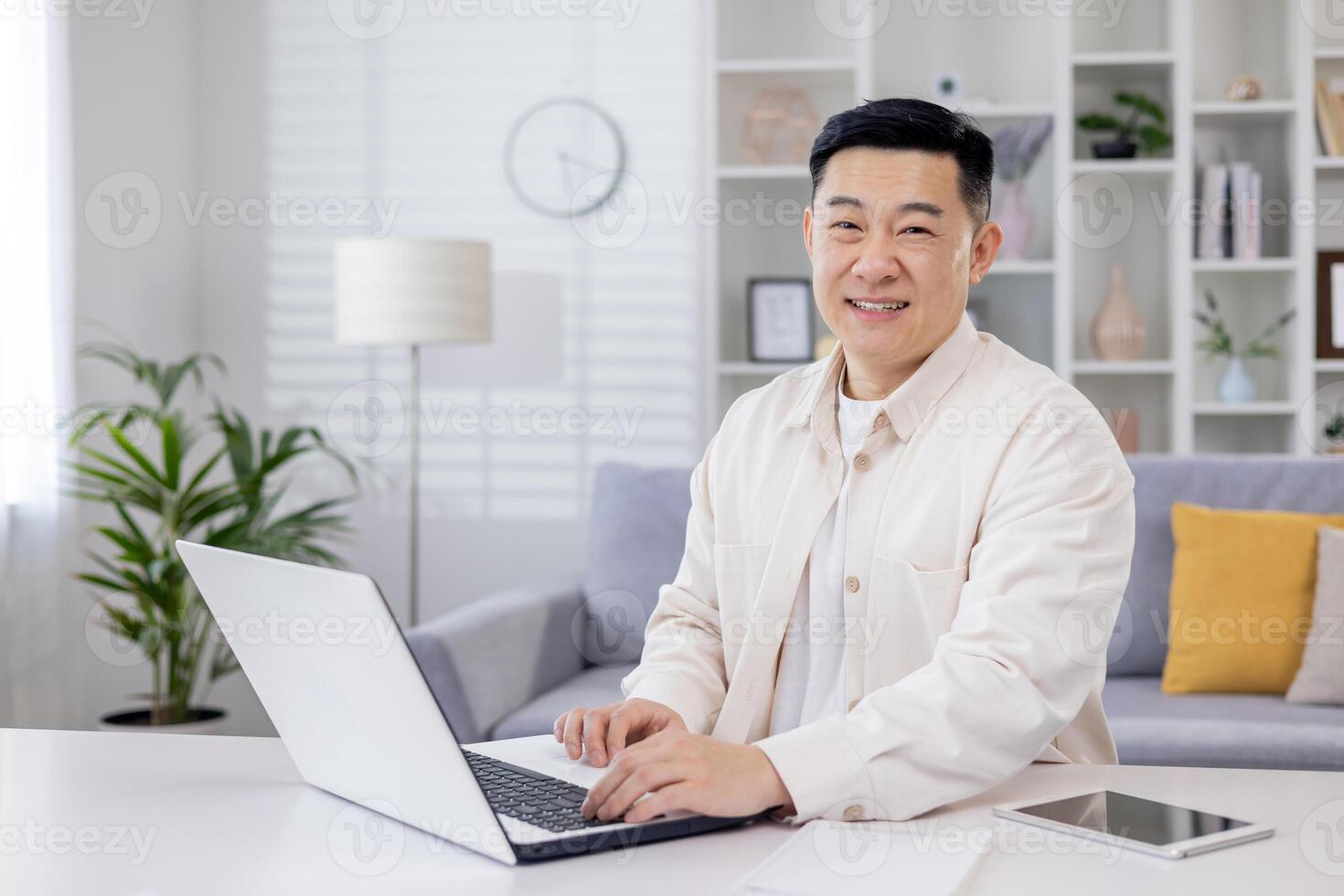 retrato de exitoso asiático programador, hombre trabajando remotamente desde hogar oficina, empresario sonriente y mirando a cámara utilizando ordenador portátil a trabajo sentado en vivo habitación a hogar. foto