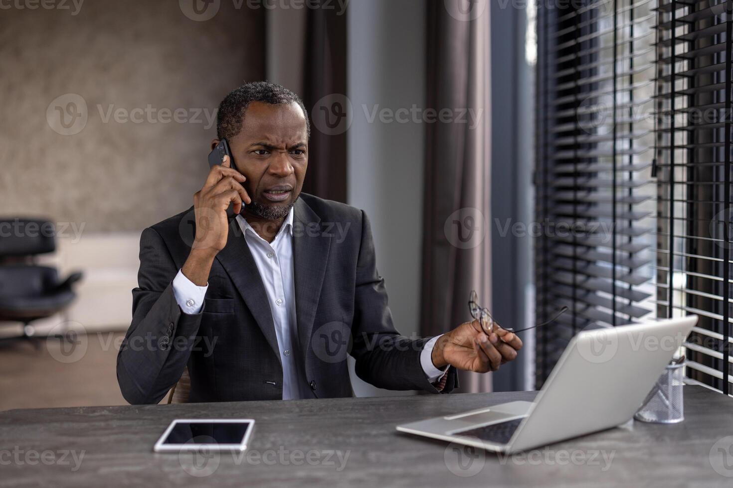 A focused African-American businessman holds glasses and talks on the phone in his modern office setting. photo