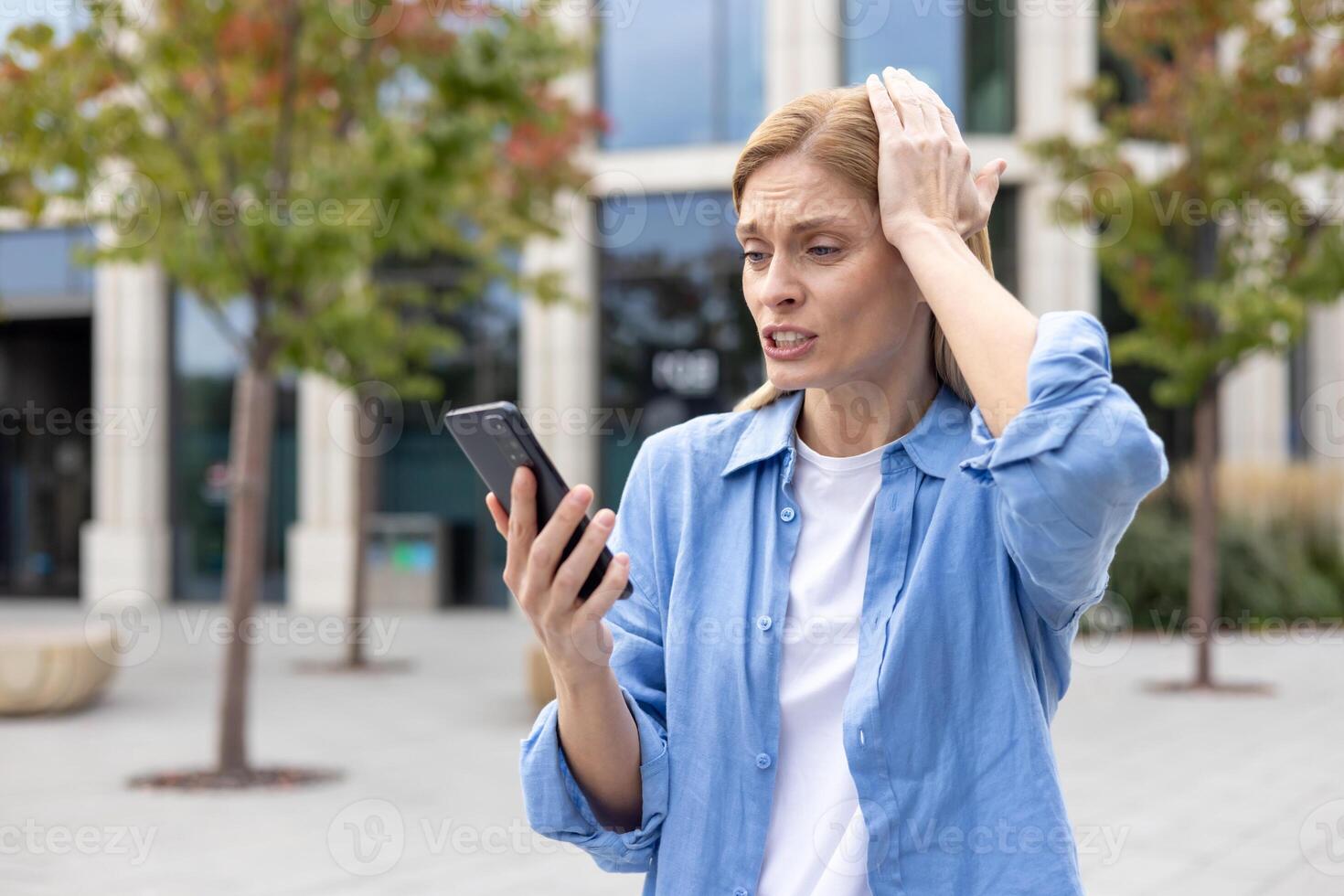 Mature woman outside office building in blue shirt sad received online message, female worker reading bad news using app on phone, viewing social media bullying and harassment. photo