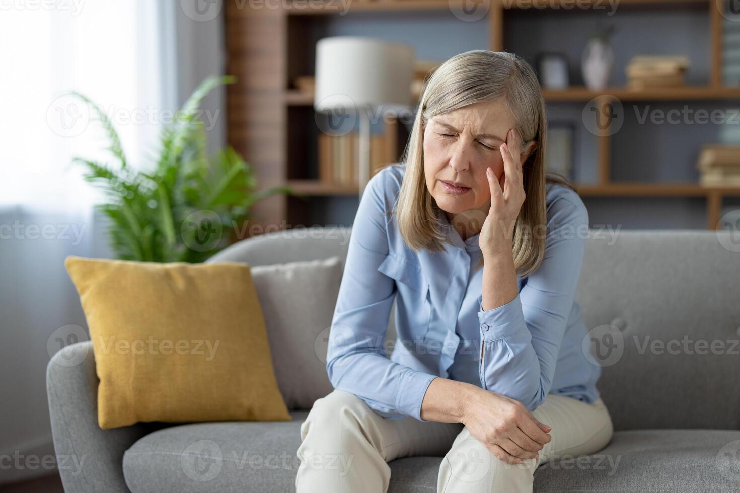 An elderly woman appears distressed and alone, sitting pensively on a couch in a well-lit living room, evoking emotions of loneliness and concern. photo