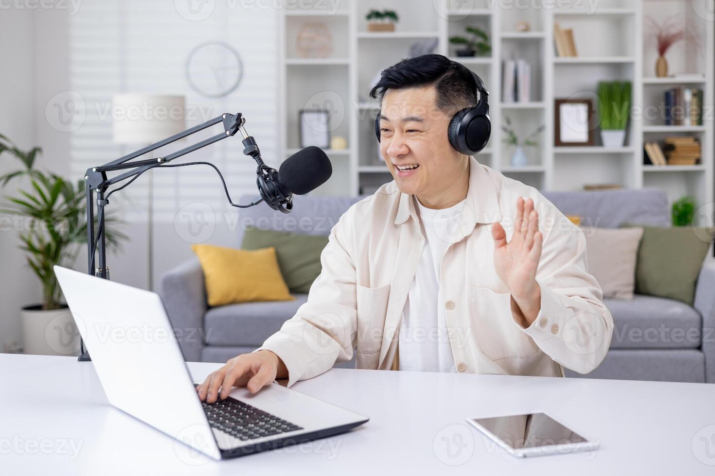 An engaging Asian man waving while recording a podcast using a laptop and microphone set up at his home office. photo