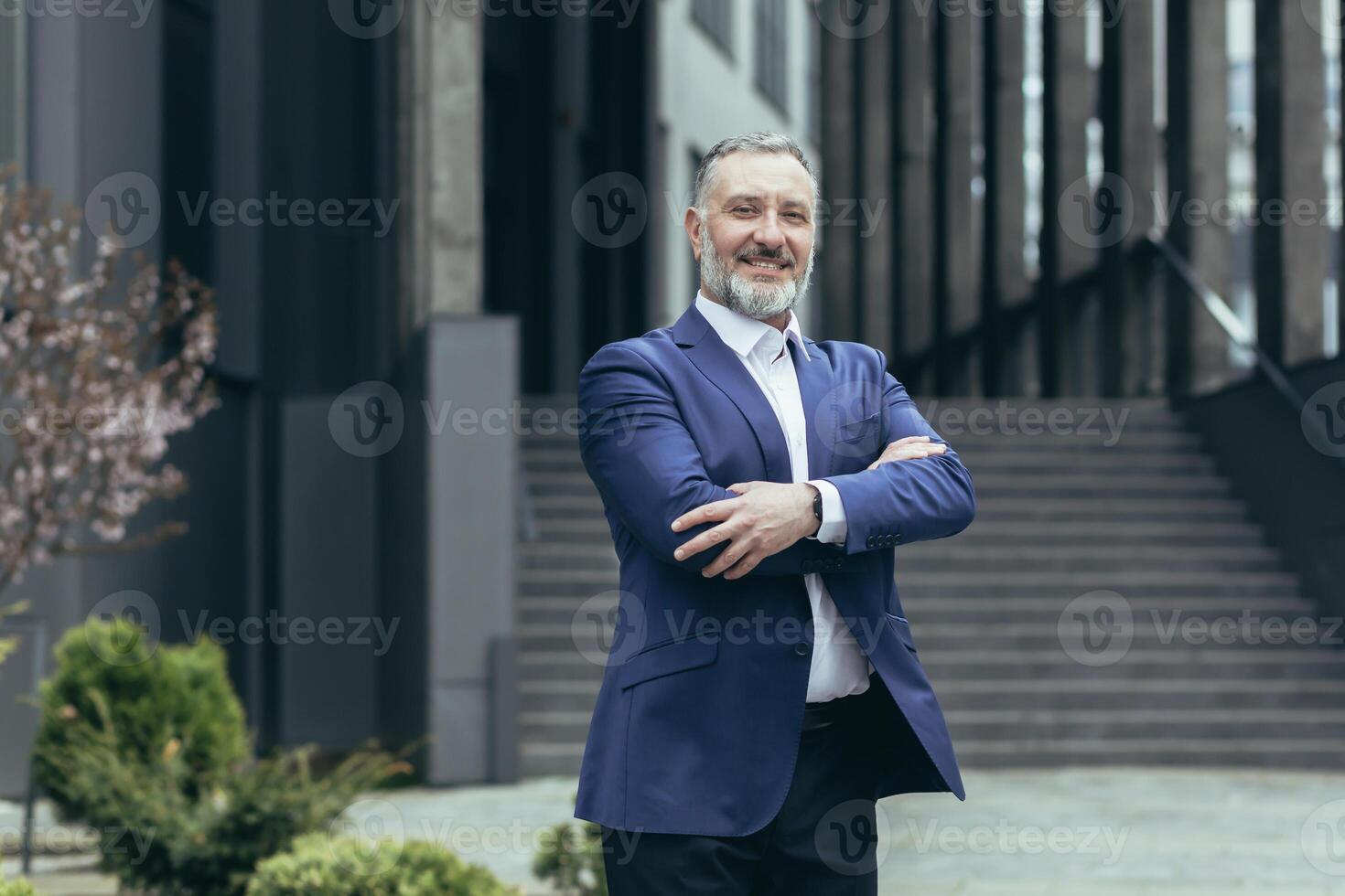 Portrait of a successful senior male businessman in a business suit standing confidently near the office center in full height, arms crossed, smiling. photo