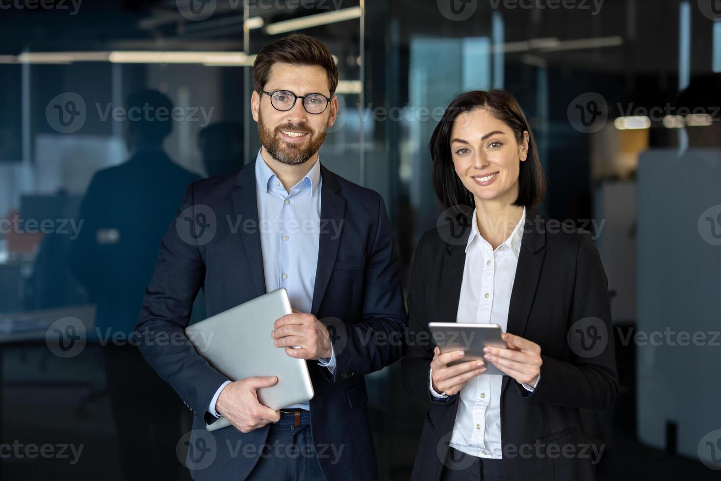 Smiling male and female in stylish suits standing together in office hall and holding digital gadgets in hands. Friendly coworkers getting ready for online meeting with boss next to boardroom. photo