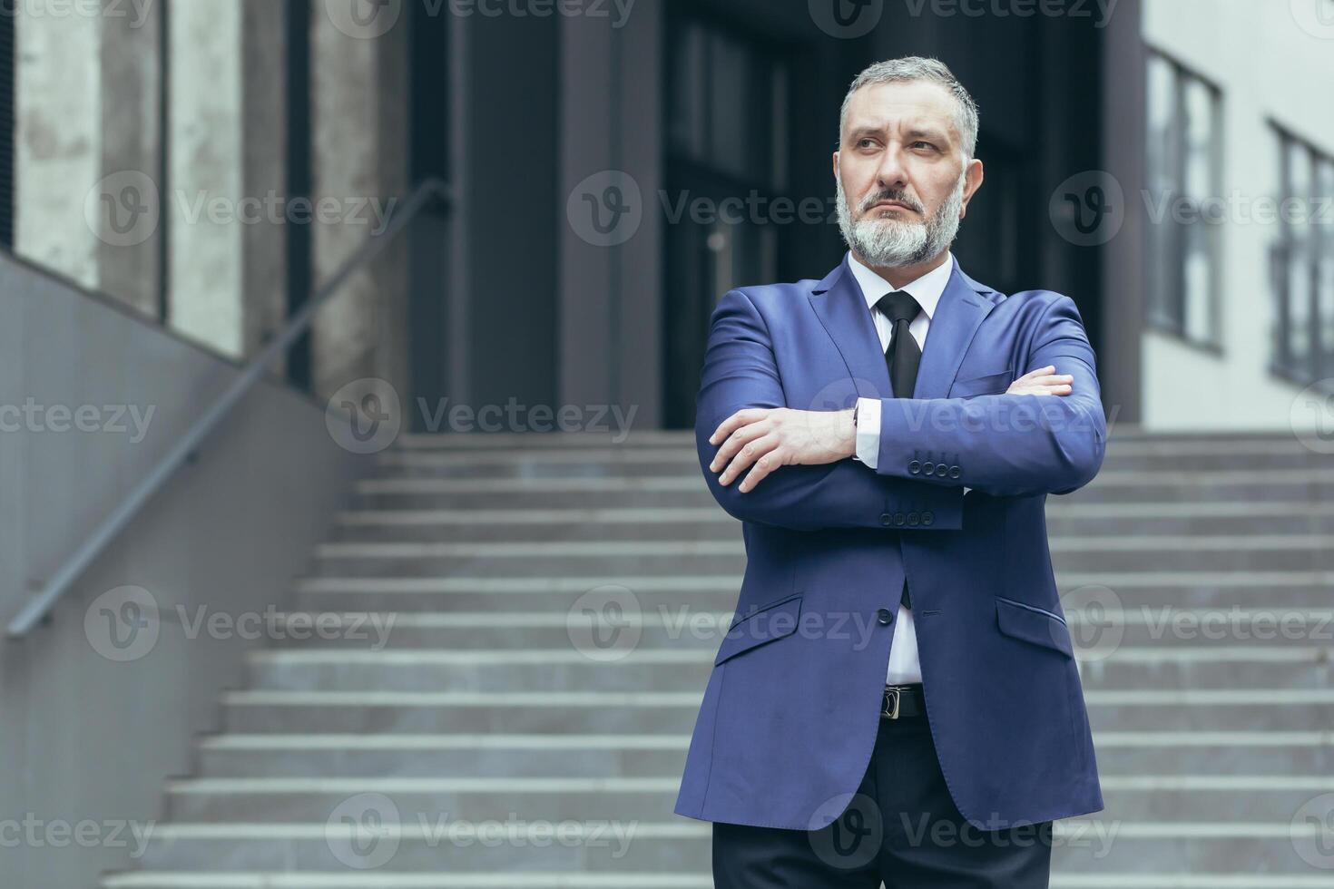 Portrait of a senior gray-haired man director, ceo on the background of the stairs of the office center. He is standing serious in a suit, arms crossed in front, looking to the side. photo