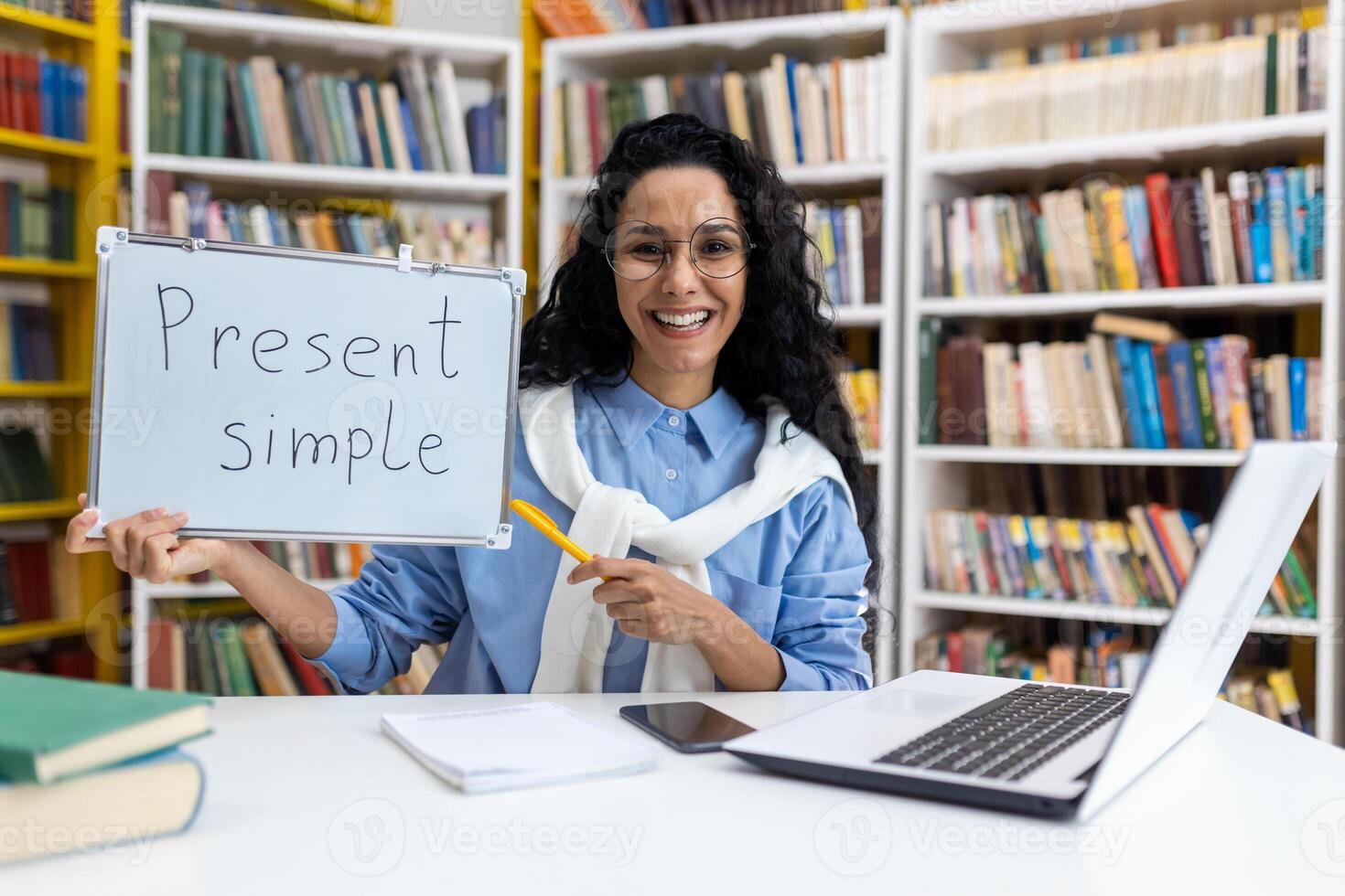 Cheerful female teacher holding a board with 'Present Simple' written on it, demonstrating English grammar in a library setting surrounded by books. photo