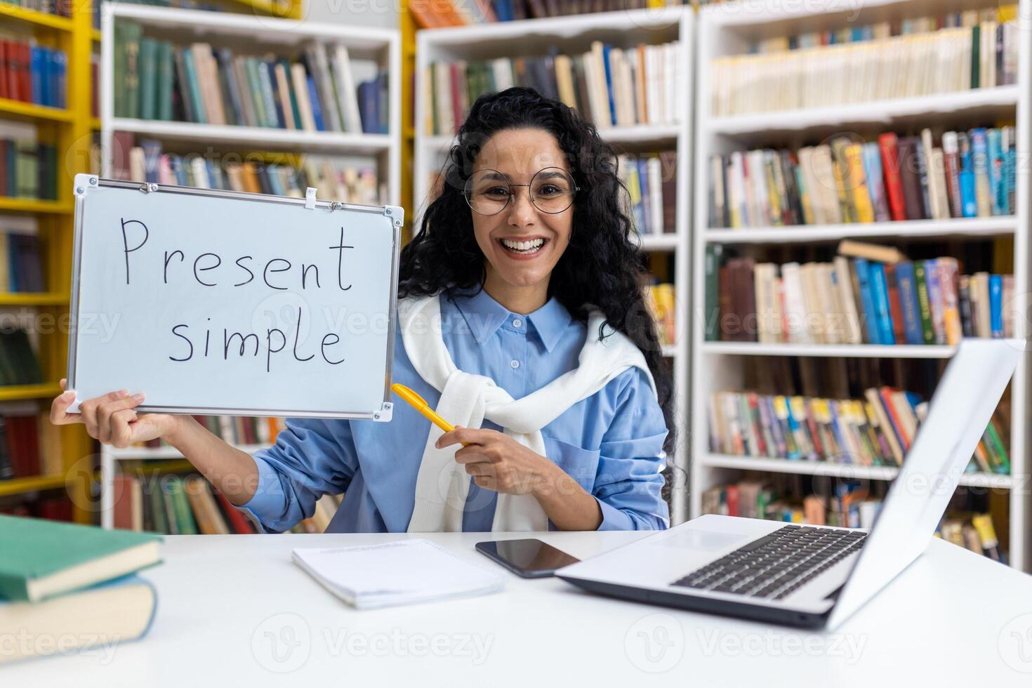 alegre hembra profesor participación un tablero con 'presente simple' escrito en él, demostrando Inglés gramática en un biblioteca ajuste rodeado por libros. foto