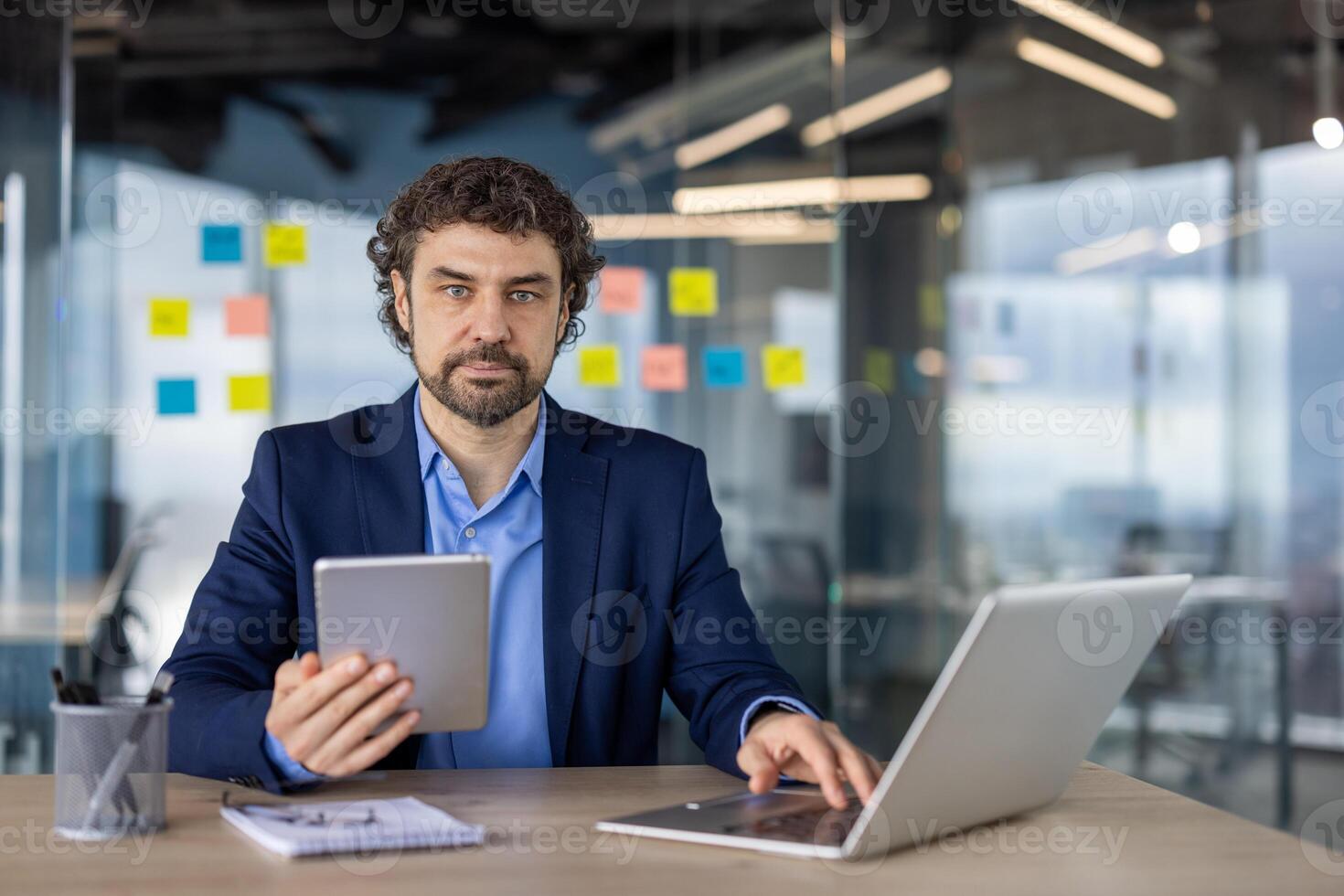 Portrait of serious confident businessman at workplace inside office, mature man looking thinkingly at camera, boss in business suit working with laptop, holding tablet computer in hands. photo